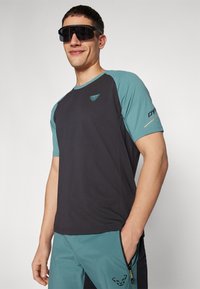 Černé a tyrkysové sportovní tričko s krátkým rukávem, kulatým výstřihem a logem na hrudi, spárované s tyrkysovými kalhotami s boční kapsou na zip.