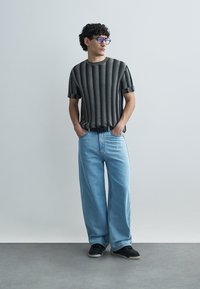 Chemise à manches courtes à rayures noires et blanches, jean large bleu clair, baskets noires, et lunettes bleues. Pose debout sur un fond neutre.