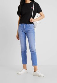 Černé bavlněné tričko s krátkým rukávem, světle modré klasické džínsy a bílé tenisky. Model drží bílou crossbody tašku.