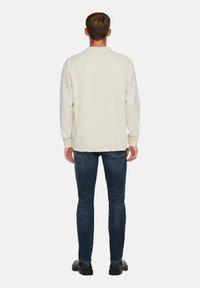 Suéter de crema claro con un ajuste relajado, mangas largas y puños de canalé, combinado con jeans azul oscuro y zapatos negros, visto desde la parte de atrás.