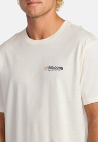 Camiseta de algodón color crema con mangas cortas. Presenta un logo de Billabong en azul marino y naranja en el lado izquierdo del pecho. Textura suave y ligera.