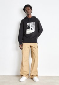 Felpa nera con stampa grafica, tasca a marsupio e cappuccio con cordino; abbinata a pantaloni cargo beige con tasche laterali e vestibilità rilassata.