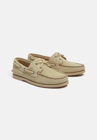 Beige mockasin-båtskor med mokassintå, snörad design, dekorativ sömnad och gummisula med beige detaljer.