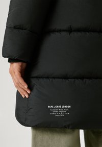 Melnā pūkainā jaka ar kvadrātveida tekstūru, kurai redzams logo un adreses detaļas baltā tekstā apakšmalā, turēta rokā.