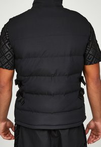 Czarna pikowana kamizelka z wysokim kołnierzem, z fakturowaną czarną tkaniną na krótkich rękawach oraz bocznymi paskami do regulacji. Matowe wykończenie.