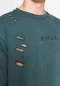 Sweatshirt oversize verde acqua in tessuto distorto, con diverse lacerazioni e un collo rotondo. Sul davanti è presente la scritta nera "MENNACE."