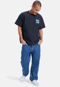 Svart kortermet t-skjorte med grafisk logo og tekst på brystet, kombinert med løse blå denimjeans og hvite sneakers.