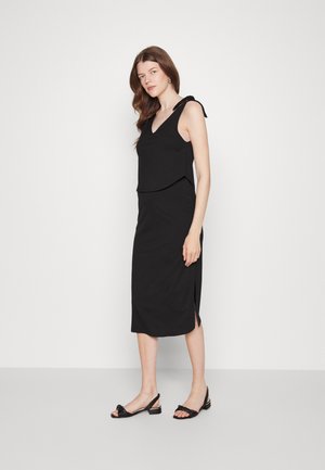Vestido preto sem mangas com decote em V, parte superior em camadas e fenda na barra. Usado com sandálias pretas de salto alto com tiras trançadas. Textura suave.