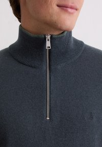 Pulover tricotat de culoare gri închis, cu un guler înalt din jerseu și un fermoar pe jumătate în față. Are un detaliu subtil cu logo pe partea stângă frontală. Textură netedă.