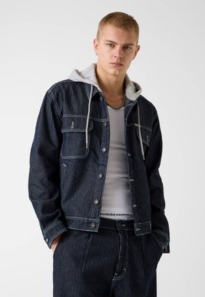 Giovane uomo che indossa una giacca e pantaloni di denim grigio scuro a righe sottili, felpa con cappuccio grigia sotto, canottiera bianca e collana di catena argento.