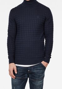 Marinblå polotröja med ett strukturerat rutmönster. Ribbstickade manschetter och nederkant, tillverkad av stickat tyg, i kombination med denimjeans.