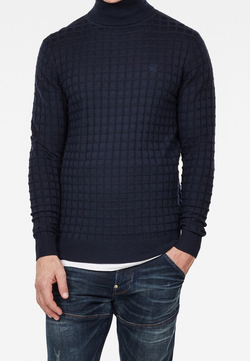 Marinblå polotröja med ett strukturerat rutmönster. Ribbstickade manschetter och nederkant, tillverkad av stickat tyg, i kombination med denimjeans.