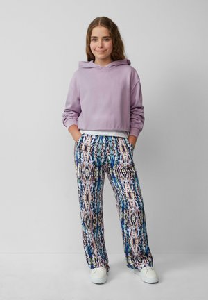 Un hoodie court lavande associé à un pantalon large coloré et à motifs avec des designs abstraits en bleu, rose et noir. Des baskets blanches.