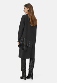 Sort kjole med lange ærmer og plisseret detalje på ryggen. Båret med knælange sorte læderstøvler. Glat tekstur, afslappet pasform.