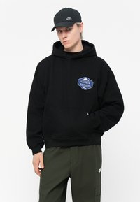 Zwarte hooded sweatshirt met een opgestikte zak aan de voorkant en een blauw embleem met de tekst "NOTES OF NOSTALGIA." Gecombineerd met olijfgroene broeken met een logo.