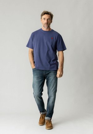 Man with short hair and beard wearing a blue t-shirt, rolled-up jeans, and brown shoes standing against a plain light background.