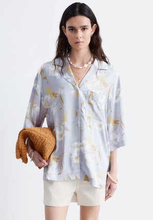 Jeune femme aux longs cheveux foncés porte un chemisier bleu clair à fleurs et un short blanc, tenant un pull en tricot marron plié dans une main.