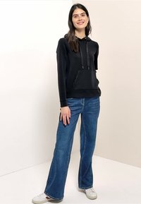 Sweat à capuche noir avec poche avant et cordons de serrage, associé à un jean évasé bleu et des baskets blanches sur un fond neutre.