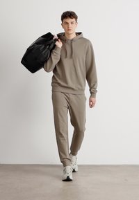 Hanorac maro deschis și pantaloni de trening asortați, fabricați dintr-un material moale. Modelul poartă o geantă mare de umăr neagră și sneakers albi.