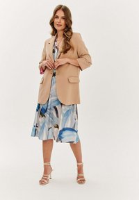 Béžový blazer s vyhrnutými rukávmi cez svetlo modré kvetinové šaty s abstraktnými vzormi štetca. Biely strappy opätky dotvárajú look.