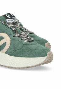 Green suede sneakers with a chunky white sole, featuring a beige logo detail and textured black-and-white laces. Highlighted stitching patterns visible.