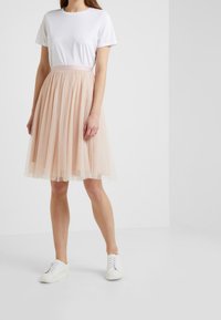 Modella che indossa una t-shirt bianca e una gonna trasparente di lunghezza ginocchio in rosa pallido con pois, abbinata a sneakers bianche, in piedi davanti a uno sfondo neutro.