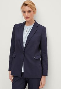 Blazer navy su misura con un solo bottone, rever a punta e due tasche frontali; abbinato a una blusa a righe azzurre sotto.