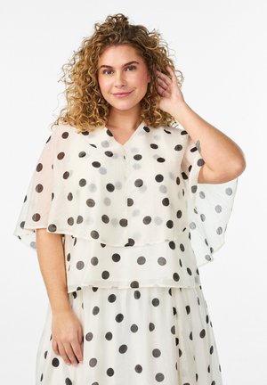 Žena s kučeravými vlasmi oblečená v bielych priesvitných šatách s bodkami a vrstveným vrchným dielom, usmievajúca sa a dotýkajúca sa vlasov na bielom pozadí.