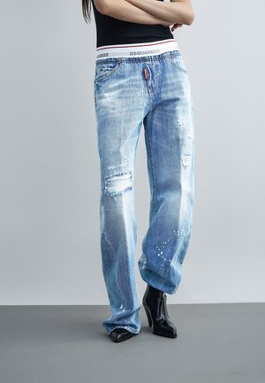 Személy világoskék, kopott farmernadrágot visel fehér DSQUARED2 elasztikus derékrésszel, fekete magassarkú csizmával és fekete ujjatlan felsővel, álló helyzetben.