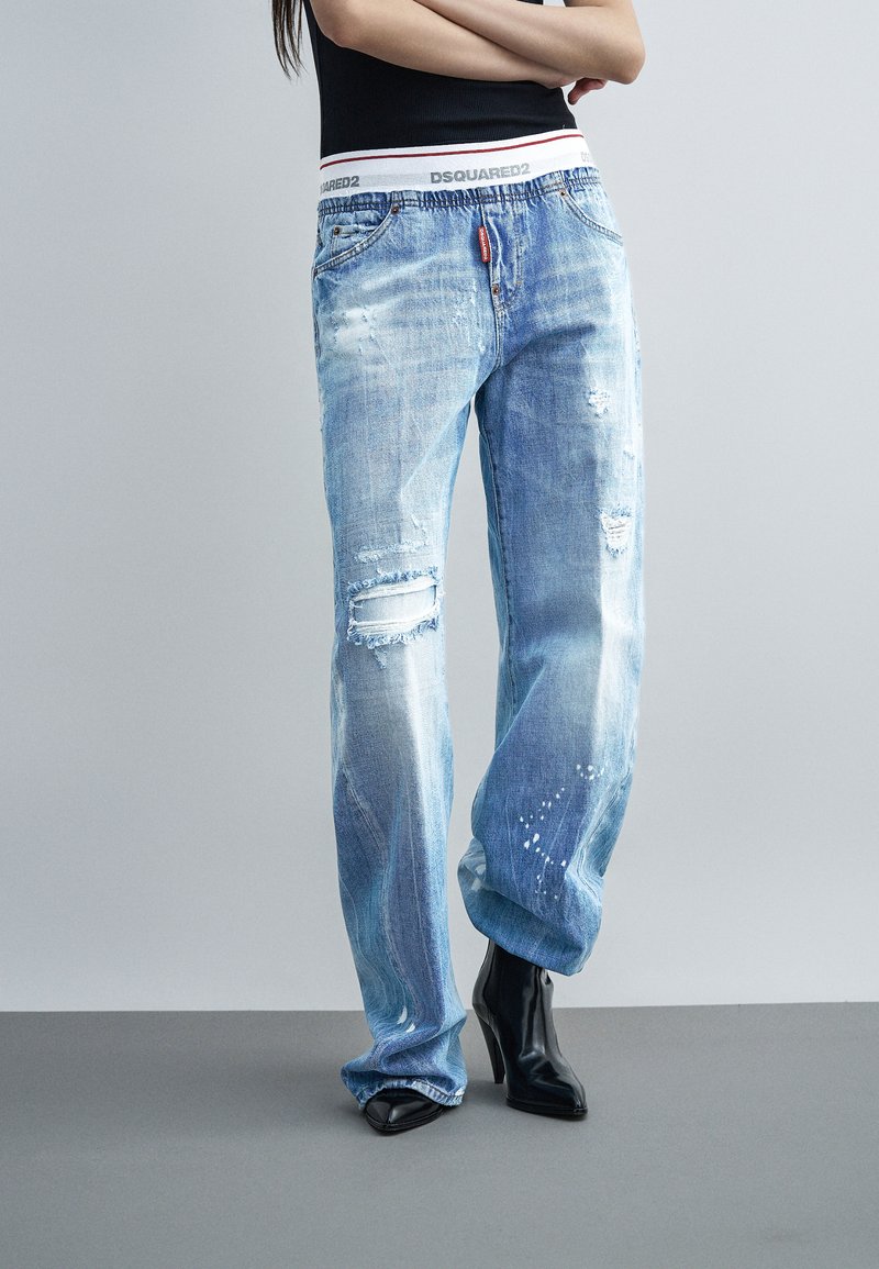 Άτομο που φοράει ανοιχτό μπλε φθαρμένο τζιν με λευκή ελαστική μέση DSQUARED2, μαύρα μπότες με τακούνι και μαύρο αμάνικο τοπ, όρθιο.