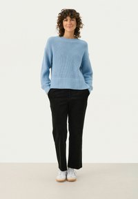 Pull en maille côtelée bleu clair avec un col rond, associé à un pantalon en velours côtelé noir et des baskets blanches sur un fond neutre.