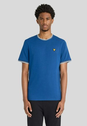 Ung mand med kort krøllet hår iført en blå t-shirt med hvid kant og et lille gult fuglelogo på brystet, står foran en grå baggrund.