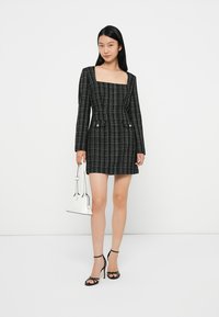 Rochie în carouri alb-negru, cu mâneci lungi, decolteu pătrat, siluetă ajustată, două buzunare frontale și detalii cu nasturi. Accesorii cu o geantă de mână albă.