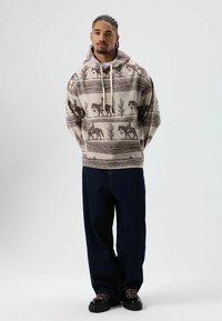 Beige en bruine hooded sweater met cactussen- en paardenpatronen, gecombineerd met donkerblauwe wijde jeans en zwarte sneakers.