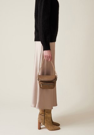 Persona che indossa una gonna midi beige, un maglione nero in peluche, stivali con tacco in tessuto tan e tiene in mano una piccola borsa abbinata con tracolla in oro.