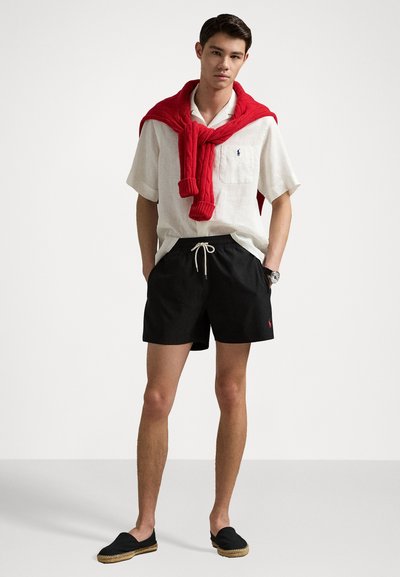 Hvit kortermet skjorte med knapper, svarte svømmebukser med snøring, rød genser knyttet rundt halsen, sorte espadrillos. Enkelt, uformelt antrekk.