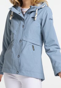 Ljusblå vattentät jacka med huva, metallknappar och dragsnören. Har en framficka och logotyp på ärmen. Slät yta.