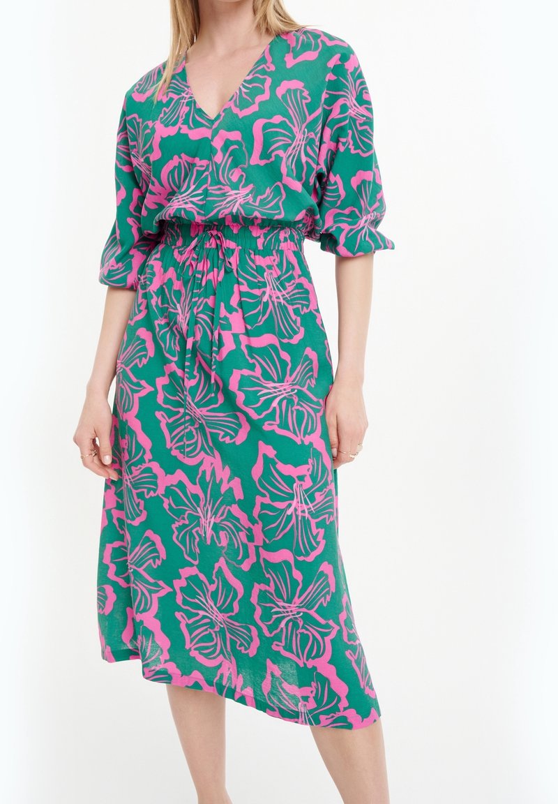 Mujer con un vestido midi verde con un patrón floral rosa, escote en V, cintura ceñida y mangas tres cuartos, sobre un fondo blanco.