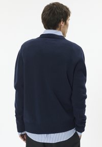 Granatowy sweter z dzianiny z prążkowanym kołnierzem i mankietami. Nosi się go na jasnoniebieską koszulę w biało-niebieskie paski, tworząc swobodny, warstwowy wygląd.