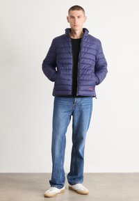 Marineblå quiltet jakke med høy krage, som har en svart glidelås og rødt logo. Kombinert med lyse jeans og hvite joggesko.
