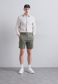 Helle beige Leinenhemd, olivgrüne Shorts, weiße Sneaker und eine schwarze Kappe. Lässiges Outfit mit lockerer Passform, das eine neutrale Farbpalette zeigt.
