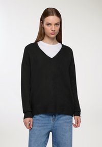 Czarny sweter z dekoltem w serek i długimi rękawami, o dzianinowej strukturze i ściągaczach mankietów, nałożony na biały T-shirt. Luźny fason z swobodną sylwetką.