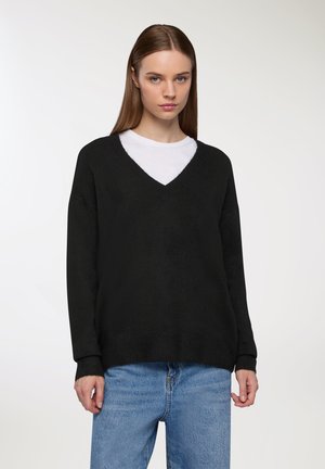 Maglione nero a V con maniche lunghe, texture a maglia e polsini a costine, indossato sopra una t-shirt bianca. Vestibilità casual con una silhouette rilassata.