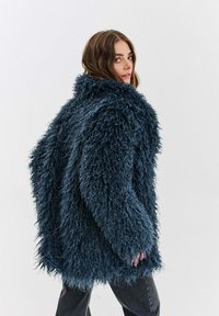 Žena v nadrozmernom tmavomodrom huňatom kabáte s textúrou, otočená bokom, pozadie svetlej farby.