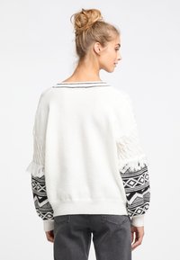 Weißer Strickpullover mit strukturiertem Körper, gemusterten schwarz-weißen Ärmeln und fransigen Manschetten. Besitzt einen kontrastierenden gestreiften Kragen.