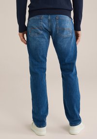 Vyras, vilkintis mėlynus džinsus ir baltus sportinius batus, pavaizduotas iš nugaros prieš neutralų fono.