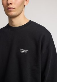 Sudadera negra con un clásico escote redondo. Presenta un logo blanco en el lado izquierdo del pecho. Textura suave y ajuste relajado.