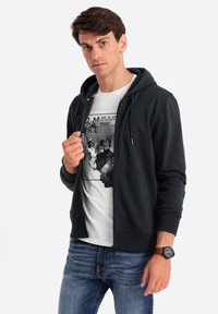 Sudadera con cremallera de color gris oscuro y bolsillo frontal, combinada con una camiseta blanca gráfica. Detalles notables incluyen cordones de contraste y un reloj.