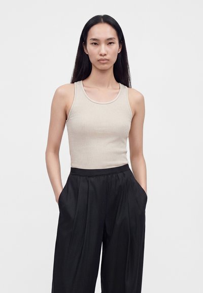 Femme aux longs cheveux noirs portant un haut côtelé beige sans manches et un pantalon noir ample taille haute, les mains dans les poches.