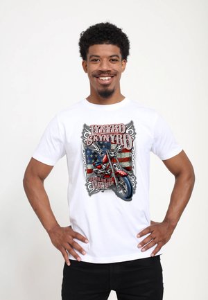 Lachende man met krullend haar die een wit Lynyrd Skynyrd T-shirt draagt met een rode motorfiets en een Amerikaanse vlag op de achtergrond.